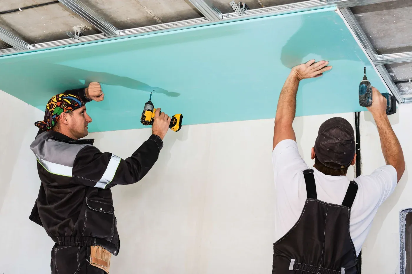 Two construction workers install a green plasterboard ceiling. Both are using drills, focused and cooperating. The scene conveys teamwork and precision.