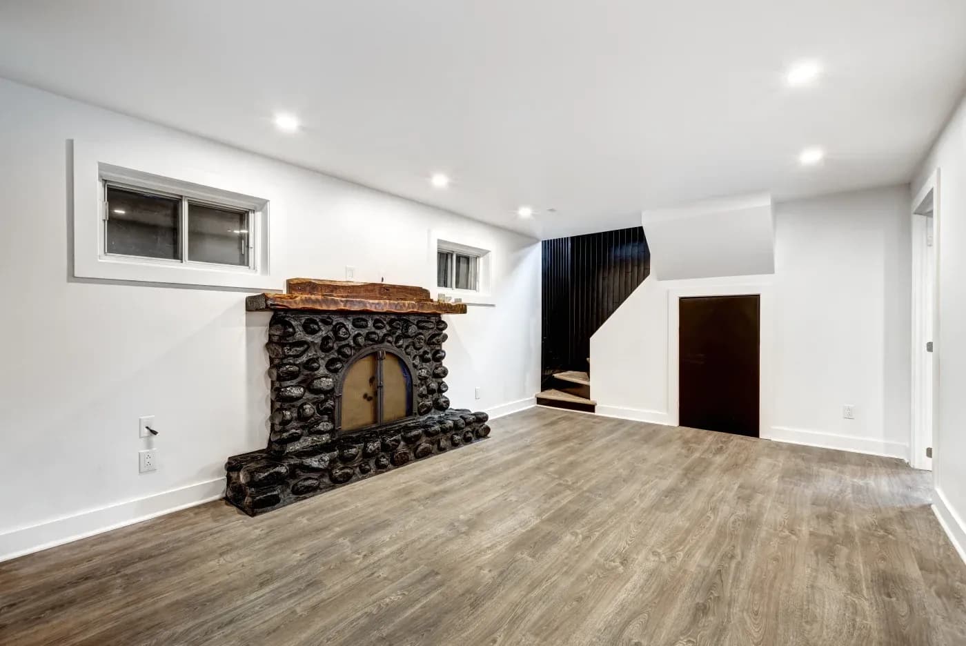 A spacious room with a rustic stone fireplace and two small windows on a white wall. It has wood flooring, recessed lighting, and a staircase.