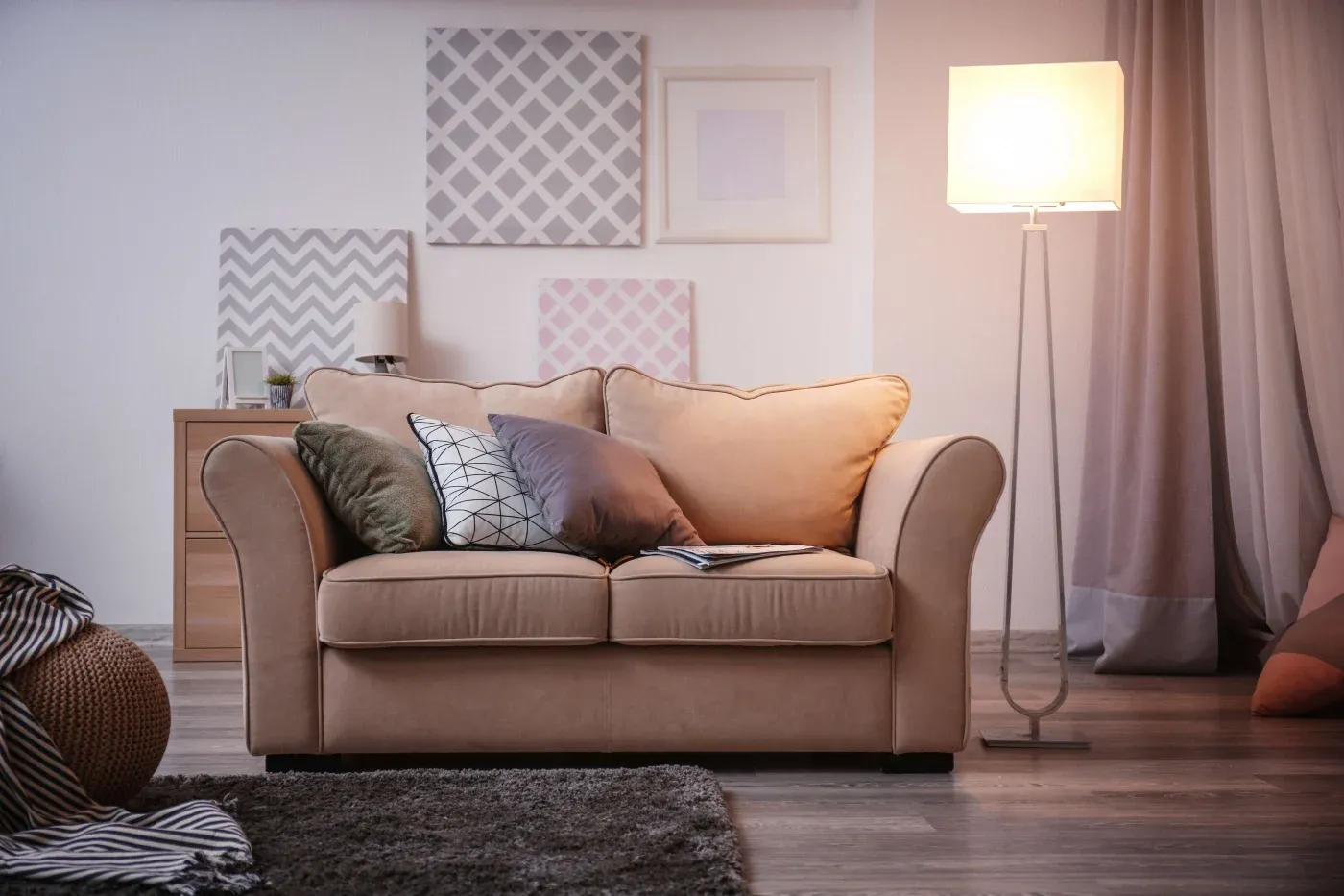 Cozy living room with a beige sofa adorned with assorted pillows. A warm lamp stands beside it, casting soft light. Geometric art decorates the white wall.