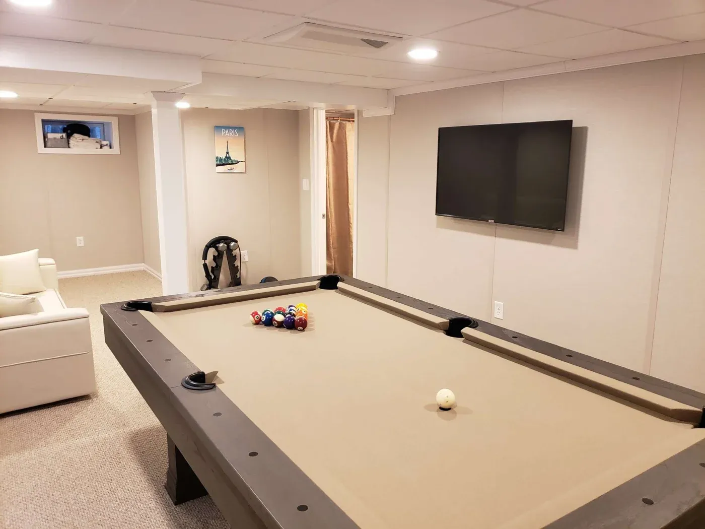 A cozy basement featuring a large billiards table with racked balls, a wall-mounted TV, a white couch, and minimal decor, creating a relaxed atmosphere.