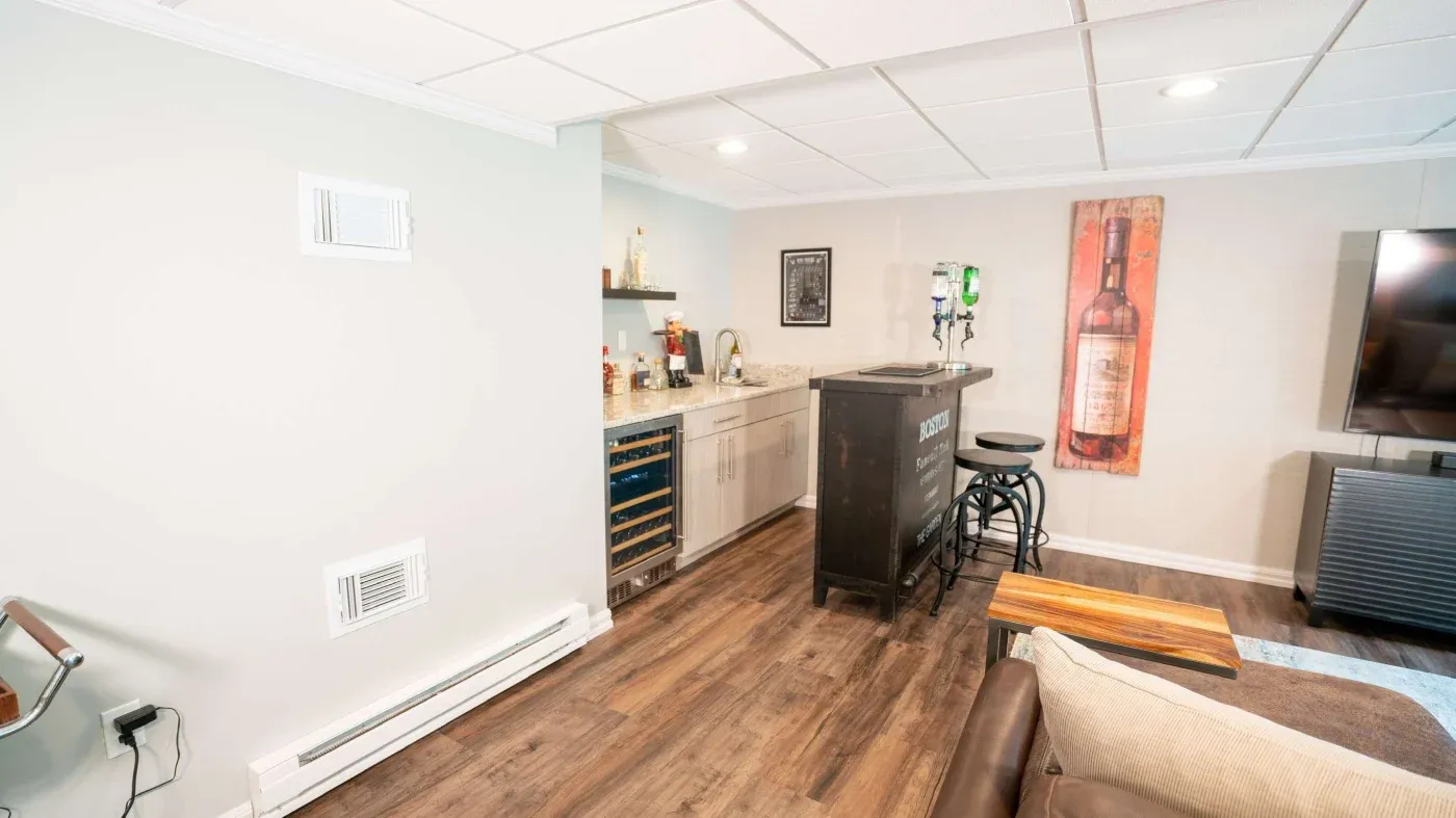 A cozy home bar with wooden floors, a dark counter, bar stools, wall shelves with liquor, and a large bottle poster. The mood is inviting and relaxed.