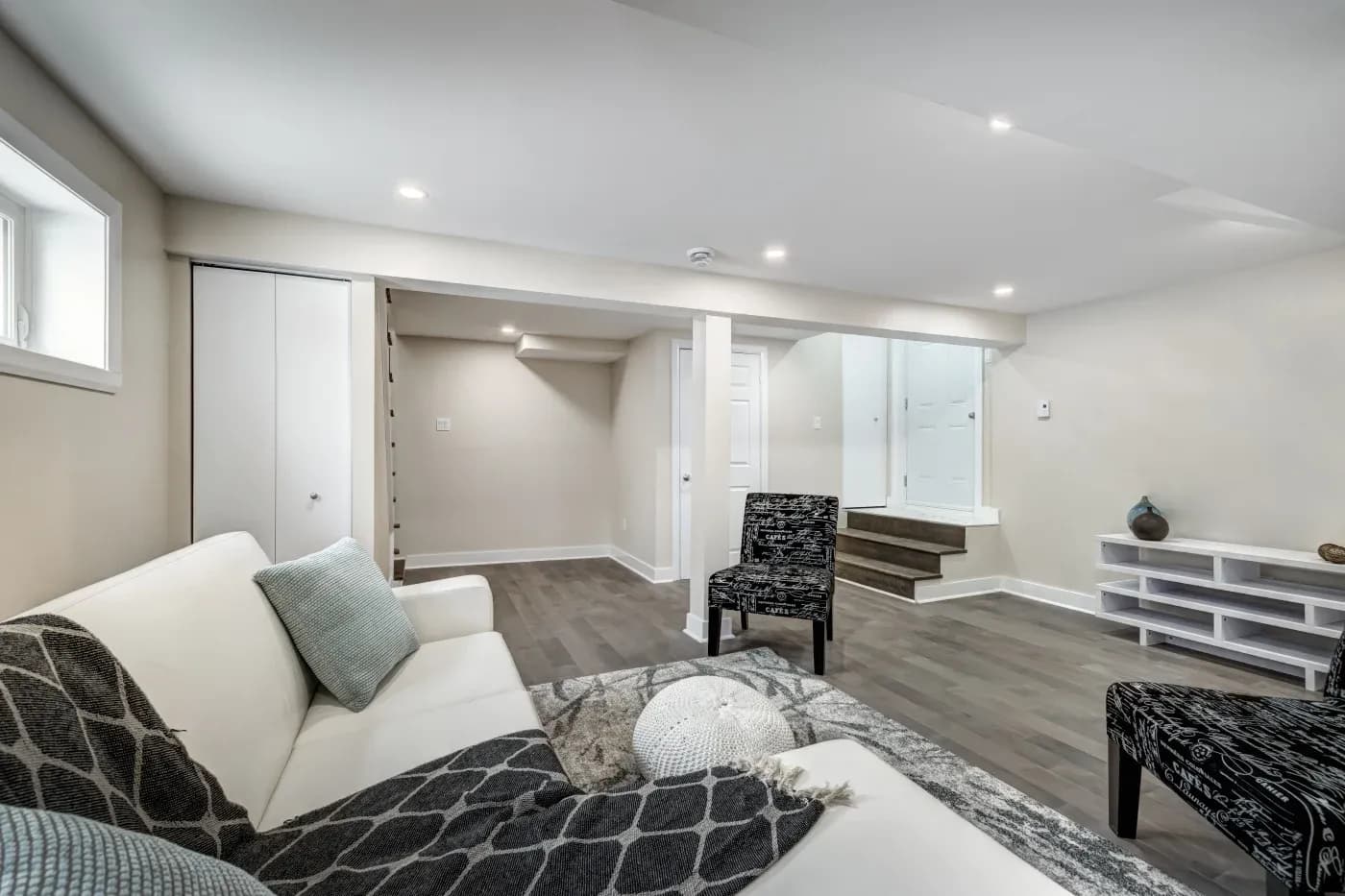 Modern basement living room with white walls, hardwood floor, and recessed lighting. Features a white couch, patterned chair, and decorative items.