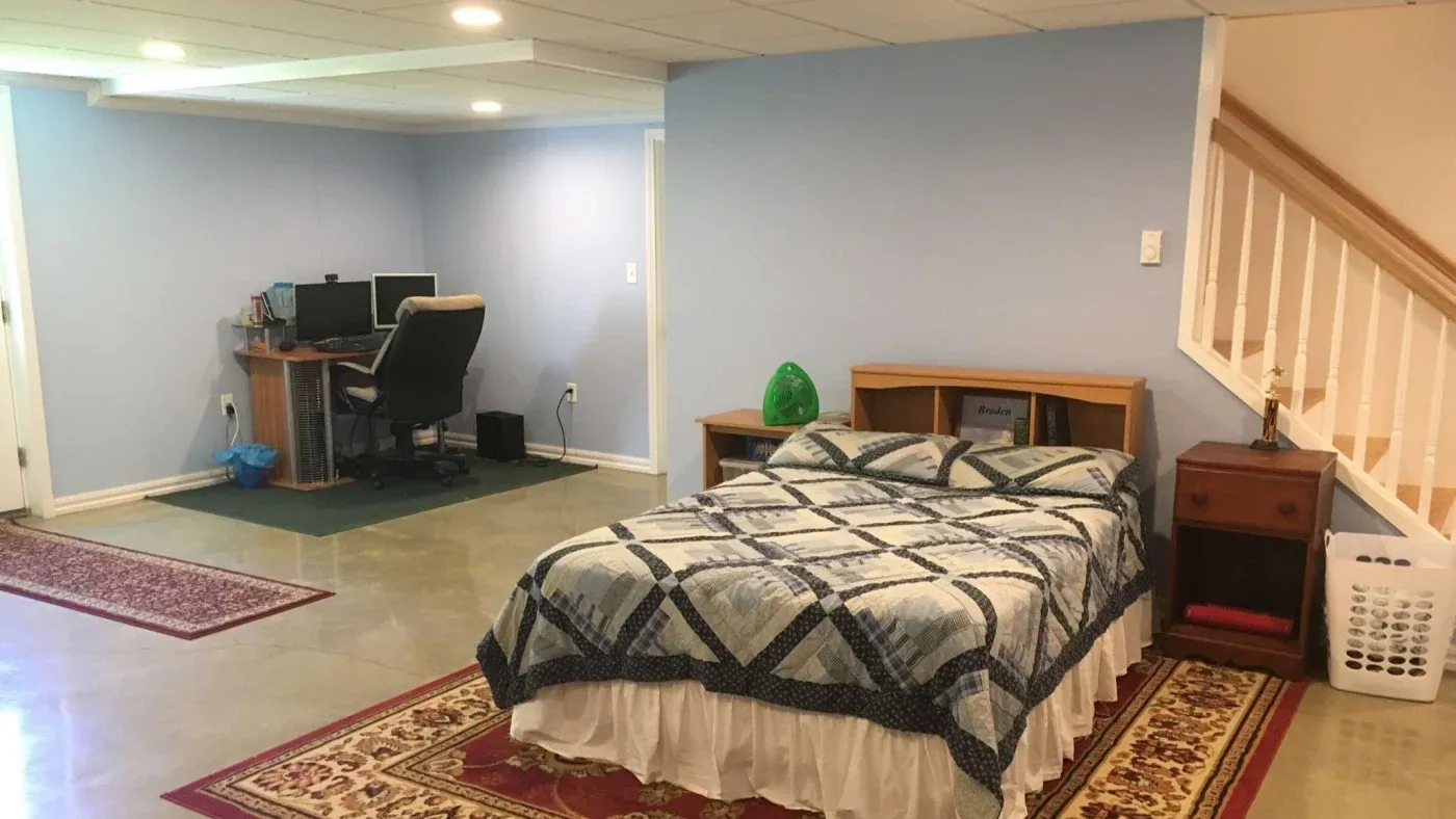 Basement room with a blue-gray wall, featuring a single bed with a patterned quilt, a wooden desk with a computer, and a laundry basket by the stairs.