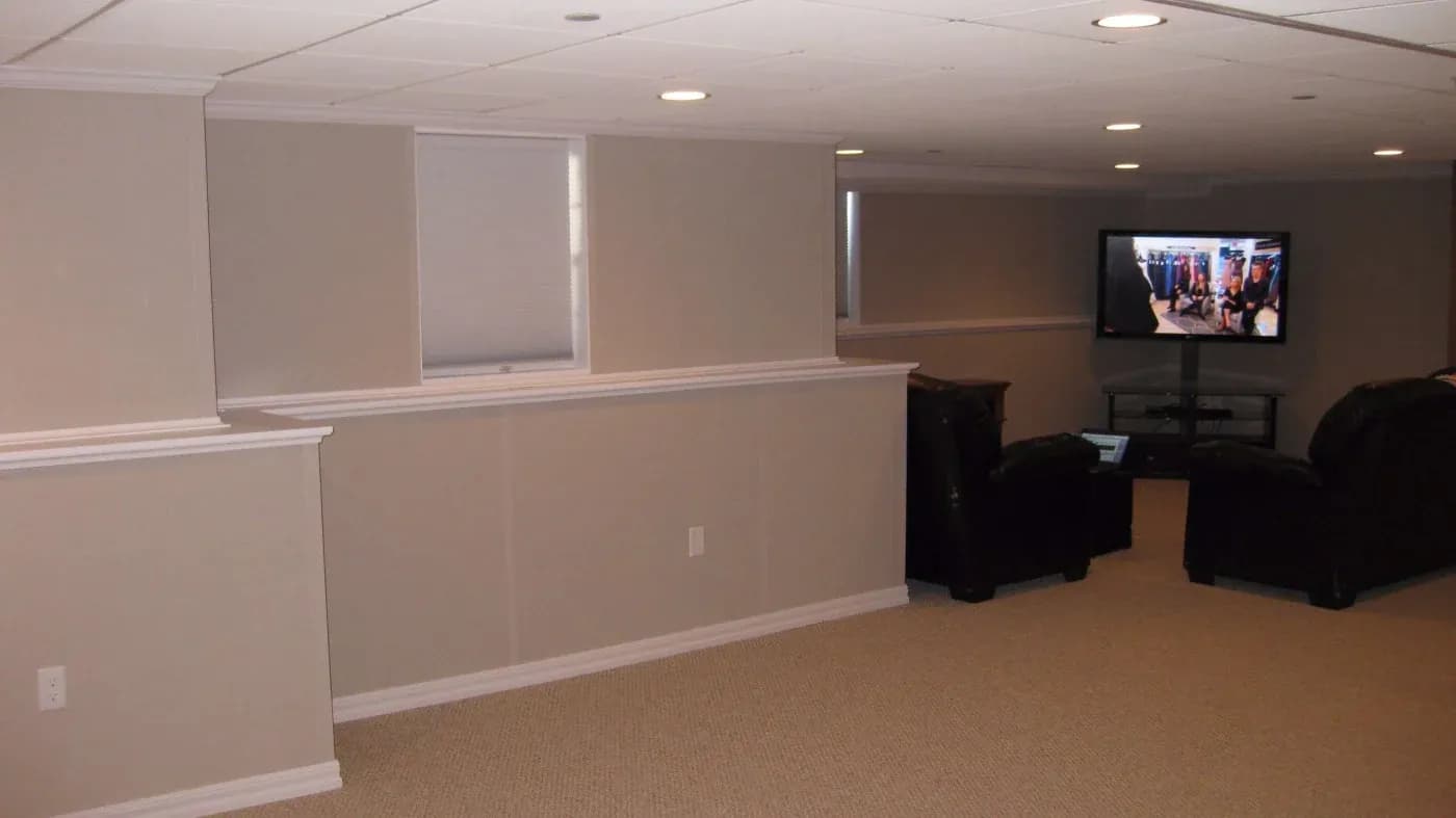 Spacious basement with beige walls, recessed lighting, and carpeted floor. Two large black leather sofas face a TV displaying a lively scene.