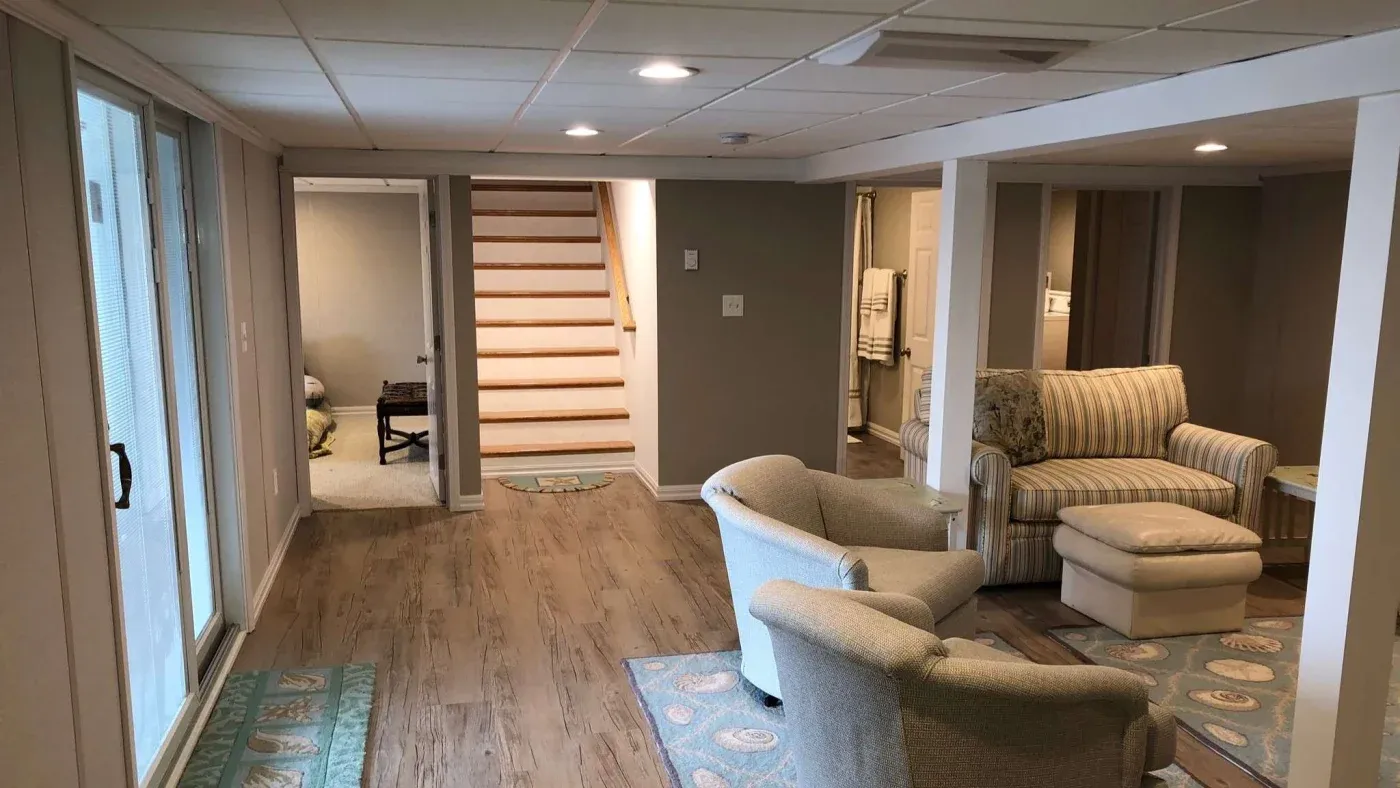 Basement living area with light wood flooring, beige walls, and recessed lighting. Features cozy armchairs, a striped sofa, and stairs leading upward.