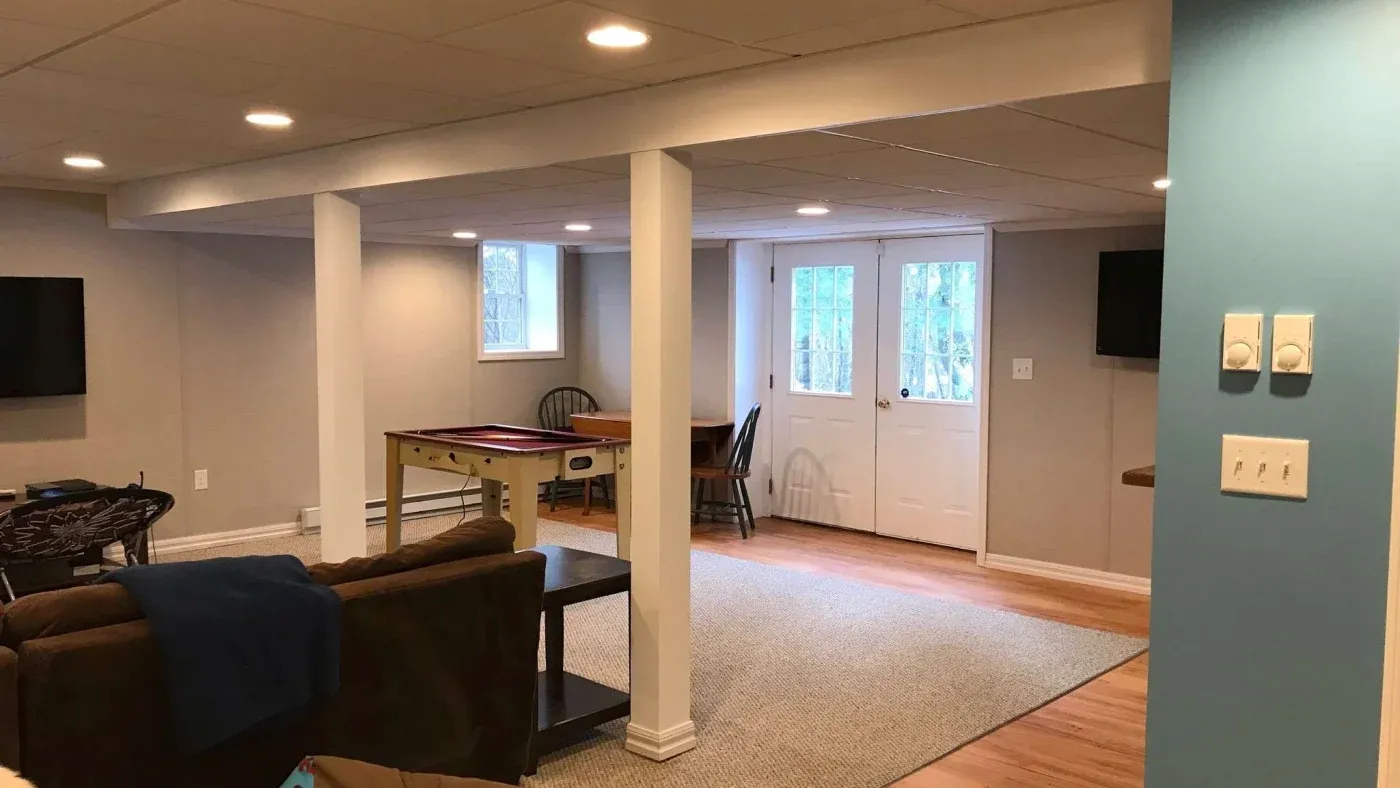 Spacious basement with beige walls features a game table, couch, and TV. Brightly lit by recessed lighting, it has a cozy and inviting atmosphere.