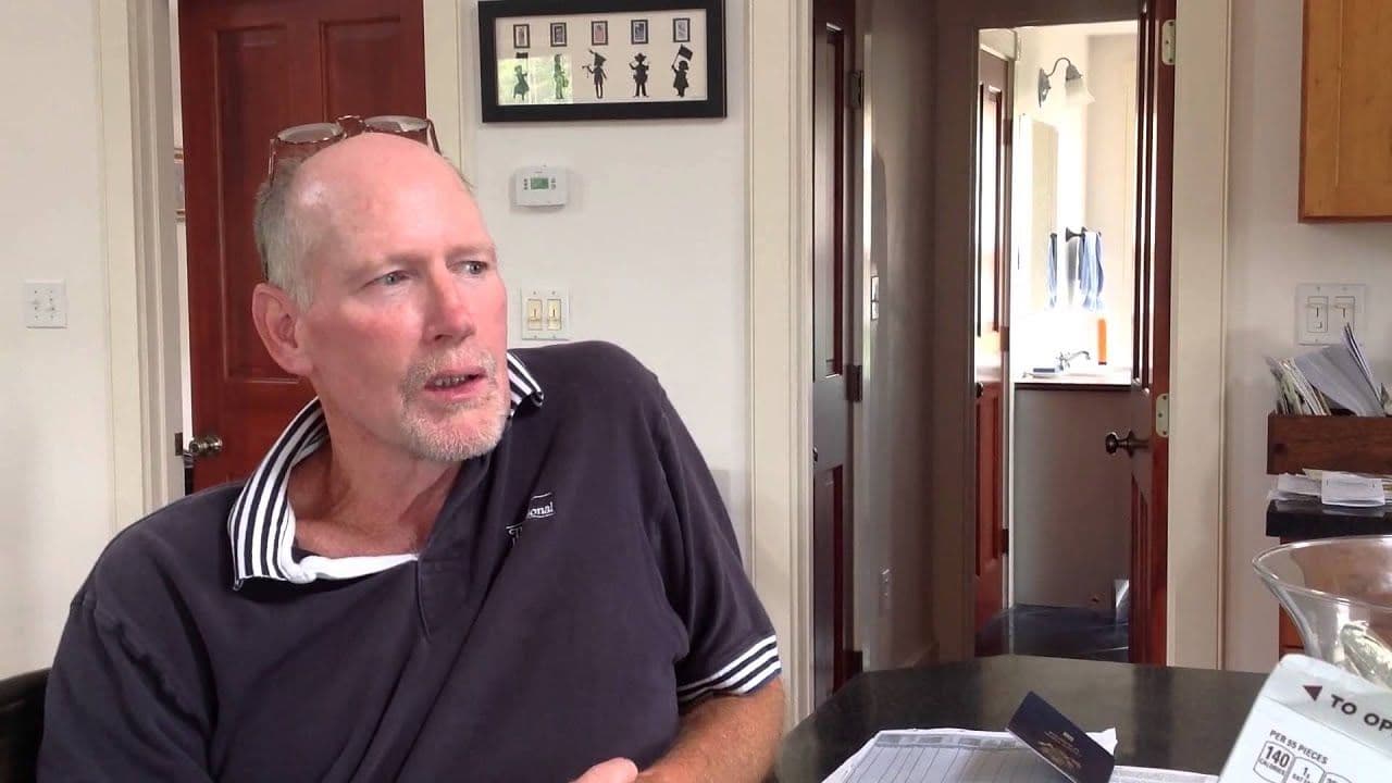 Man sitting at a kitchen table, wearing a dark polo shirt with striped collar. He appears thoughtful, with glasses on his head. Background shows open doorways.