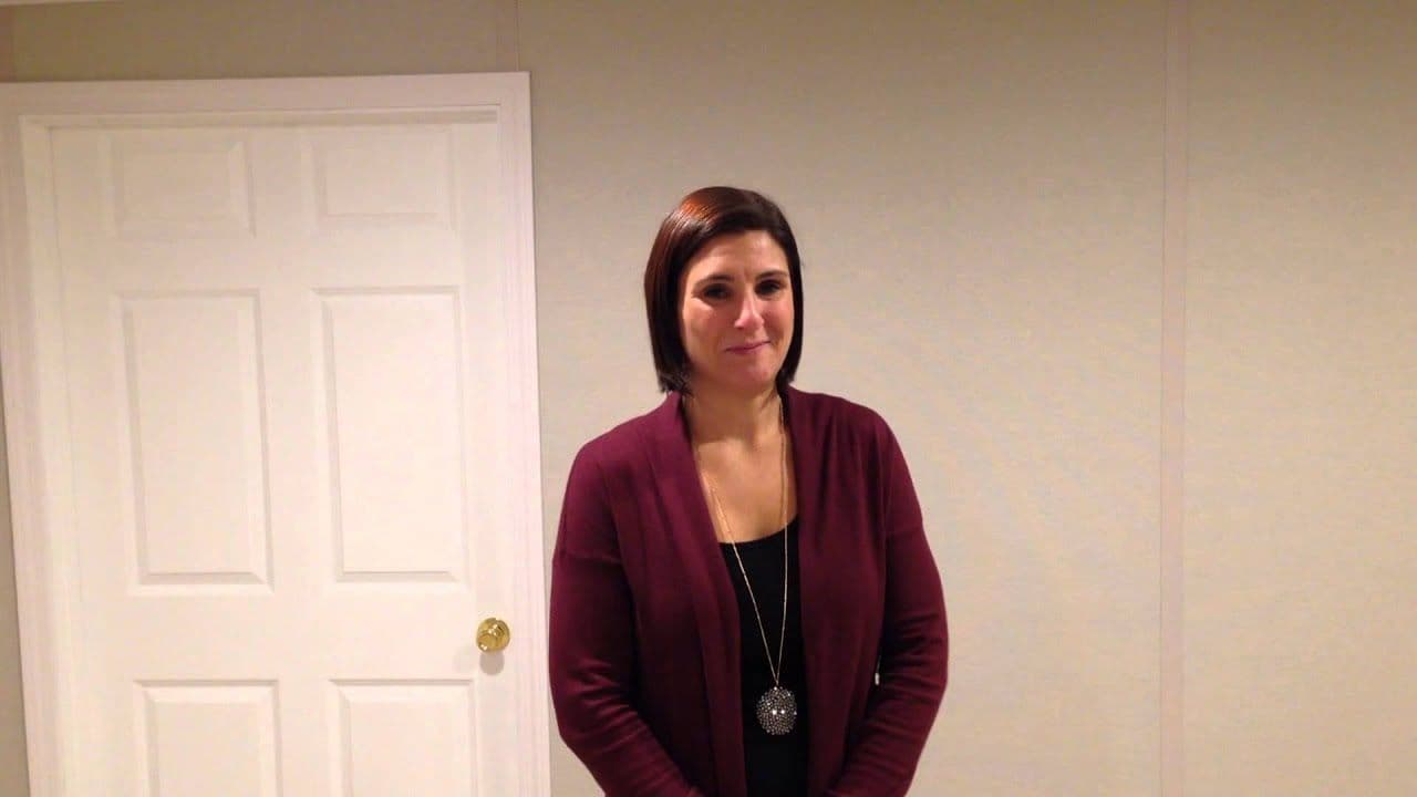 A woman in a maroon cardigan stands in front of a plain wall and a closed white door. She has short brown hair and wears a long necklace. The setting appears calm and neutral.