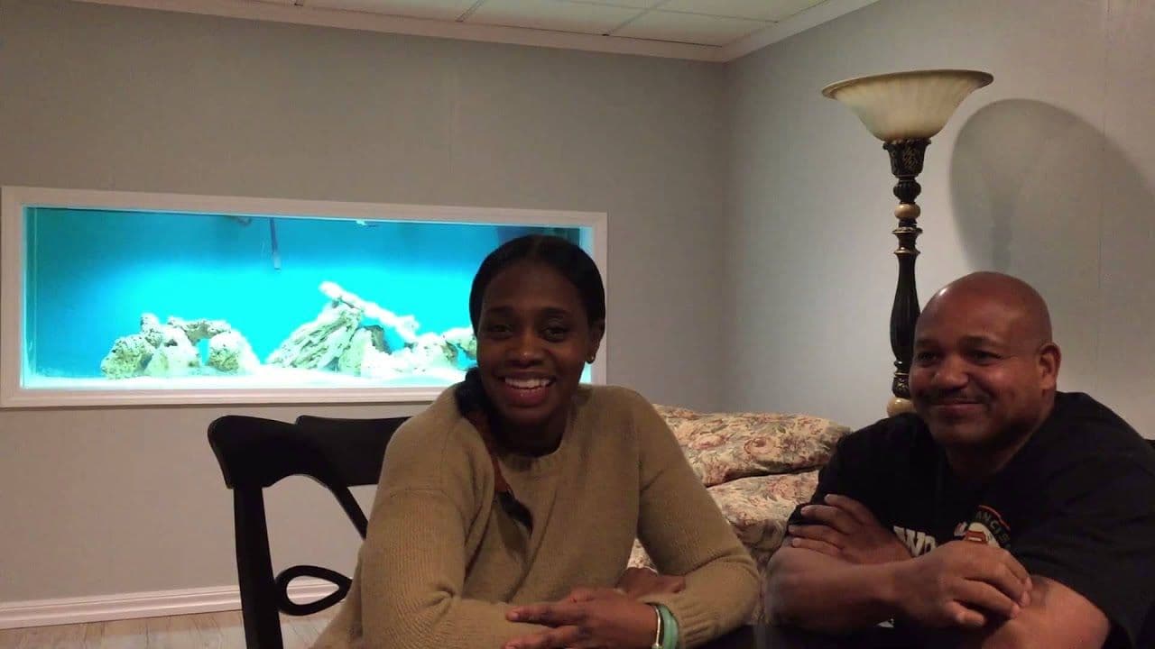 A smiling woman and man sit at a table in a cozy room. Behind them is a large aquarium with coral and rocks, creating a serene atmosphere.