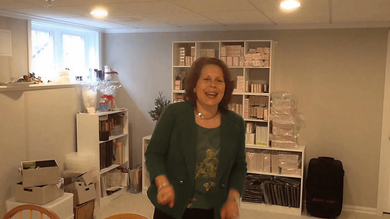 Woman in a green jacket smiles and gestures in a well-lit room with shelves of organized boxes and books, creating a cheerful and welcoming atmosphere.