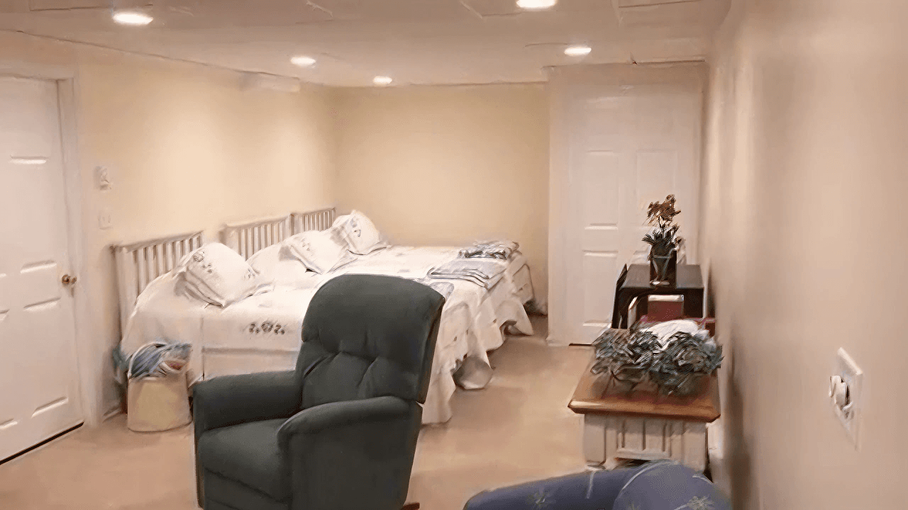 Basement room with two twin beds adorned with white linens against a beige wall. Two armchairs provide seating, and a small table holds potted plants, creating a cozy atmosphere.