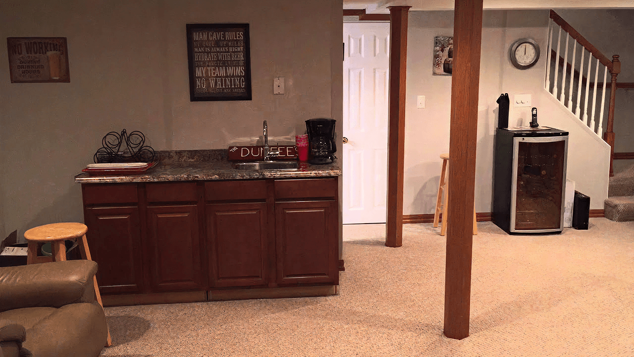 A cozy basement with a small kitchenette featuring wooden cabinets, a sink, and a coffee maker. A mini fridge stands nearby. A clock, stairs, and neutral walls create a homey vibe.