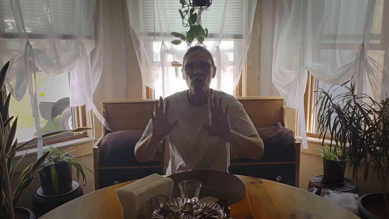 A person is sitting at a wooden table with a decorative centerpiece, surrounded by plants and sheer curtains, in a softly lit room, giving a warm, serene atmosphere.