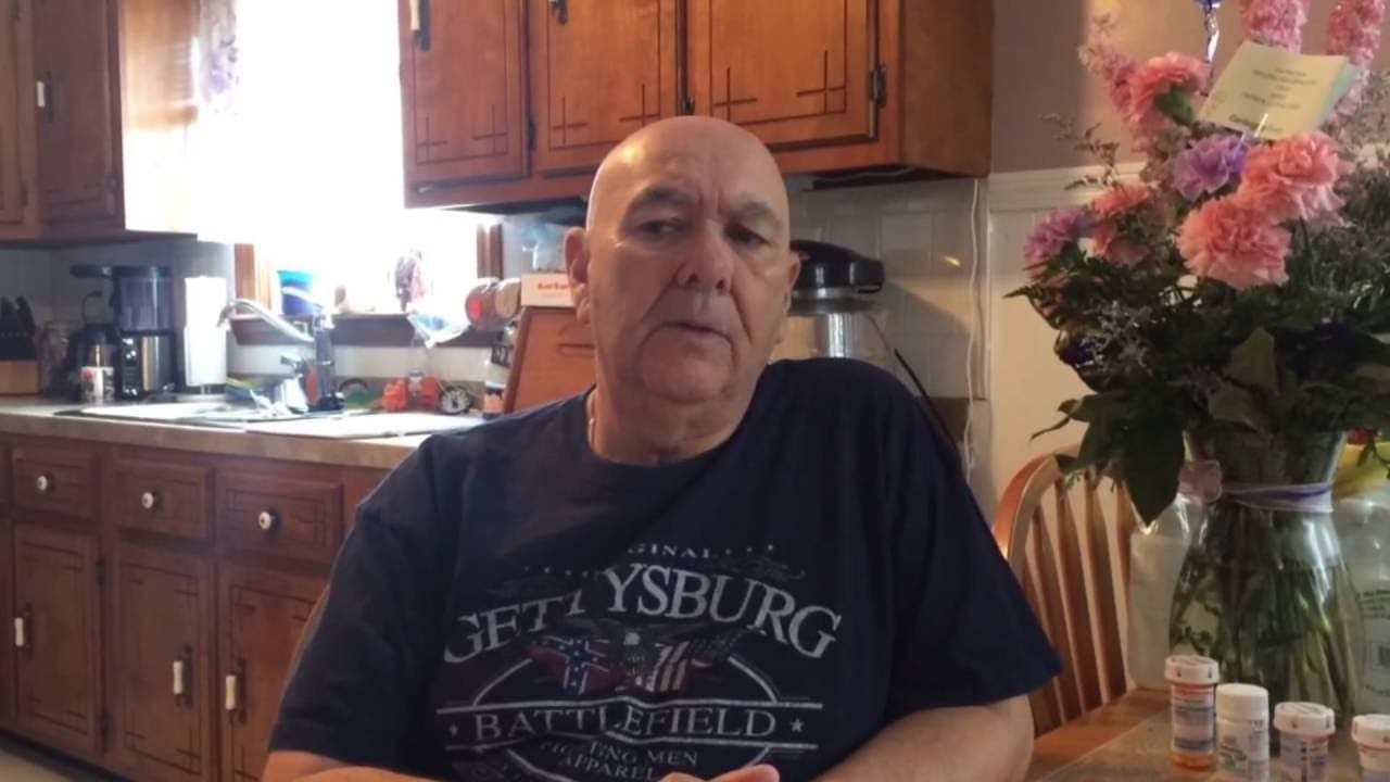 A man sits at a kitchen table wearing a Gettysburg t-shirt, surrounded by wooden cabinets and flowers. The setting feels casual and homely.