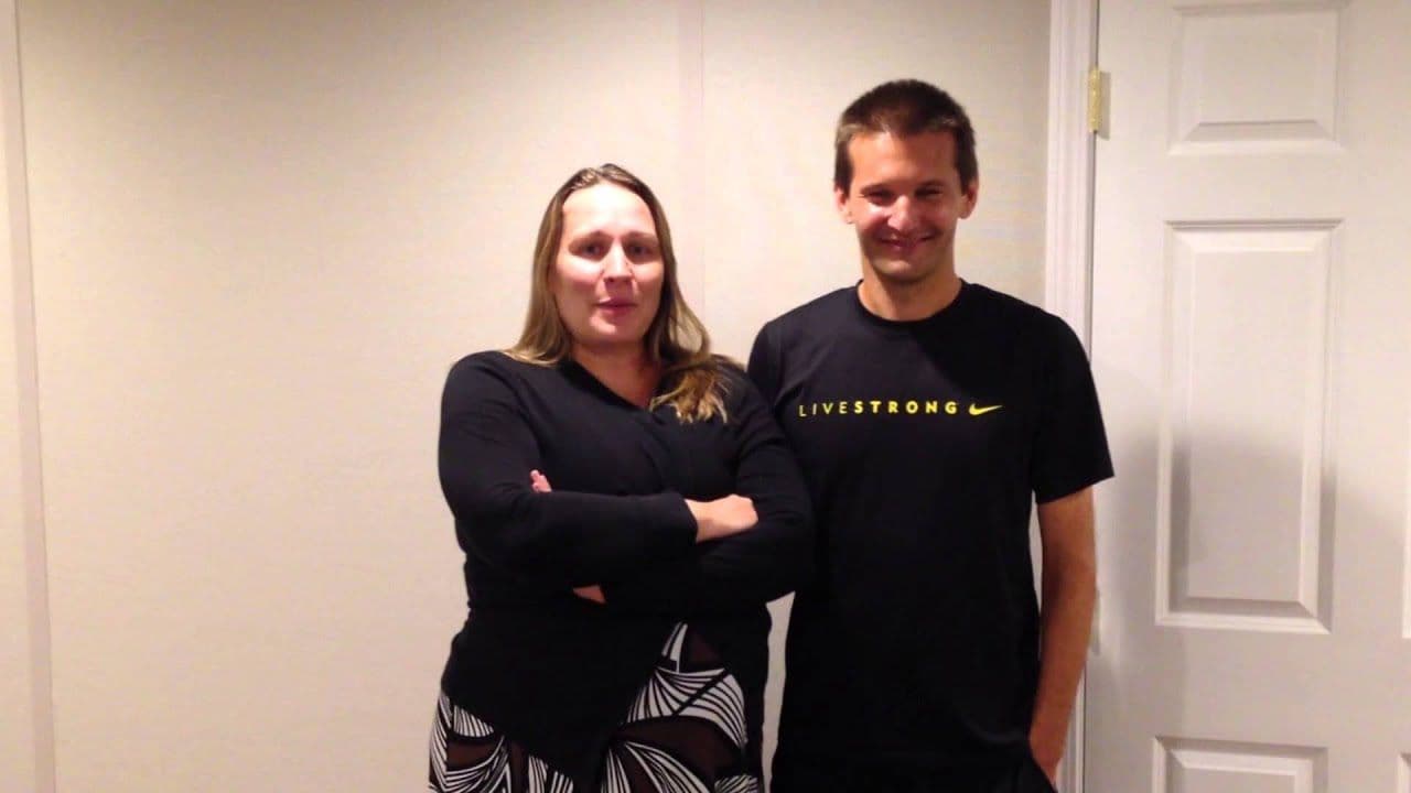 A man and woman stand side by side indoors, smiling. The woman, in a long-sleeve black top and patterned skirt, crosses her arms. The man, wearing a black "LIVESTRONG" shirt, has his hands in his pockets. They appear relaxed and content.
