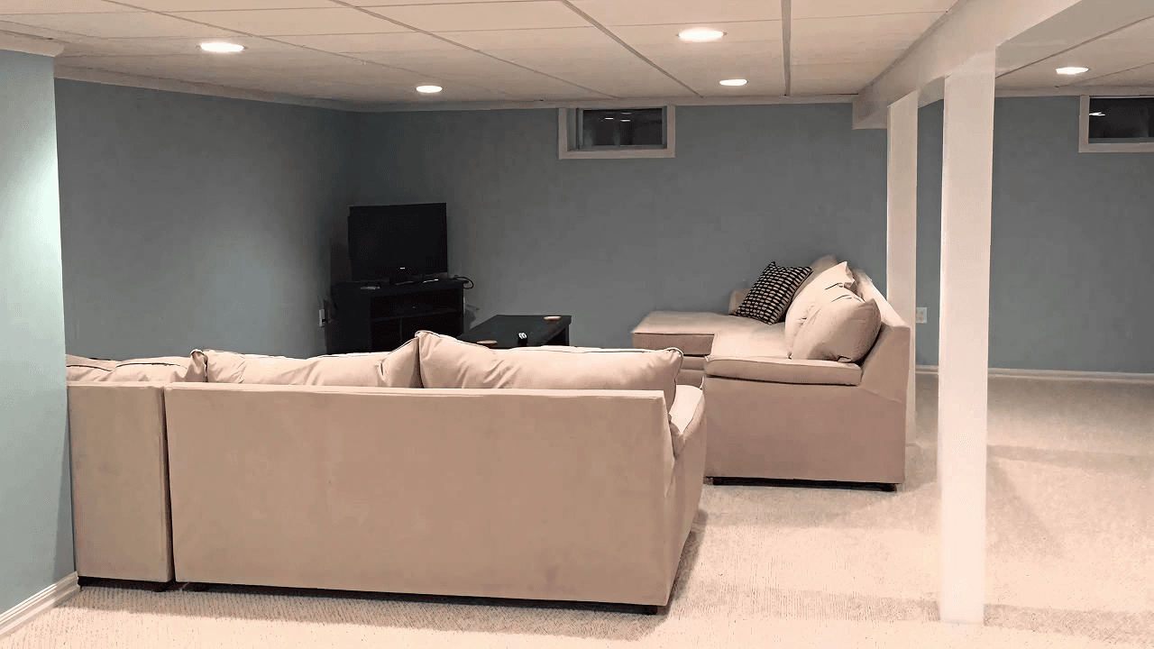 Spacious basement with beige sectional sofas on light carpet, blue walls, and recessed lighting. A TV is on a black stand, creating a cozy ambiance.