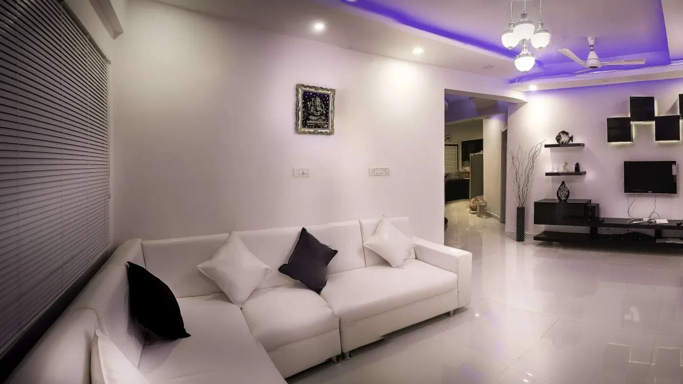 Modern living room with white sectional sofa and cushions, sleek black TV unit, contemporary decor, blue LED ceiling lights, and glossy flooring.