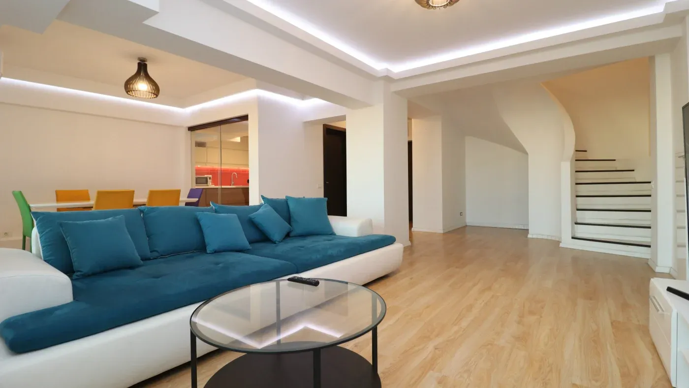 Spacious living room with a modern design featuring a large blue sectional sofa, round glass coffee table, dining area with colorful chairs, and curved staircase.