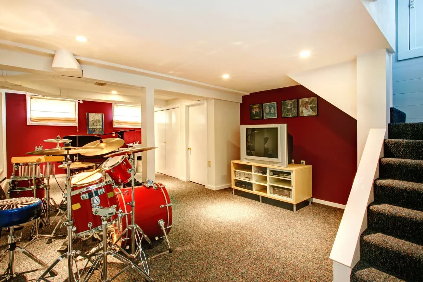 A cozy basement featuring a red drum set on a carpeted floor, a TV on a wooden stand against a maroon wall, and soft lighting creating a warm ambiance.