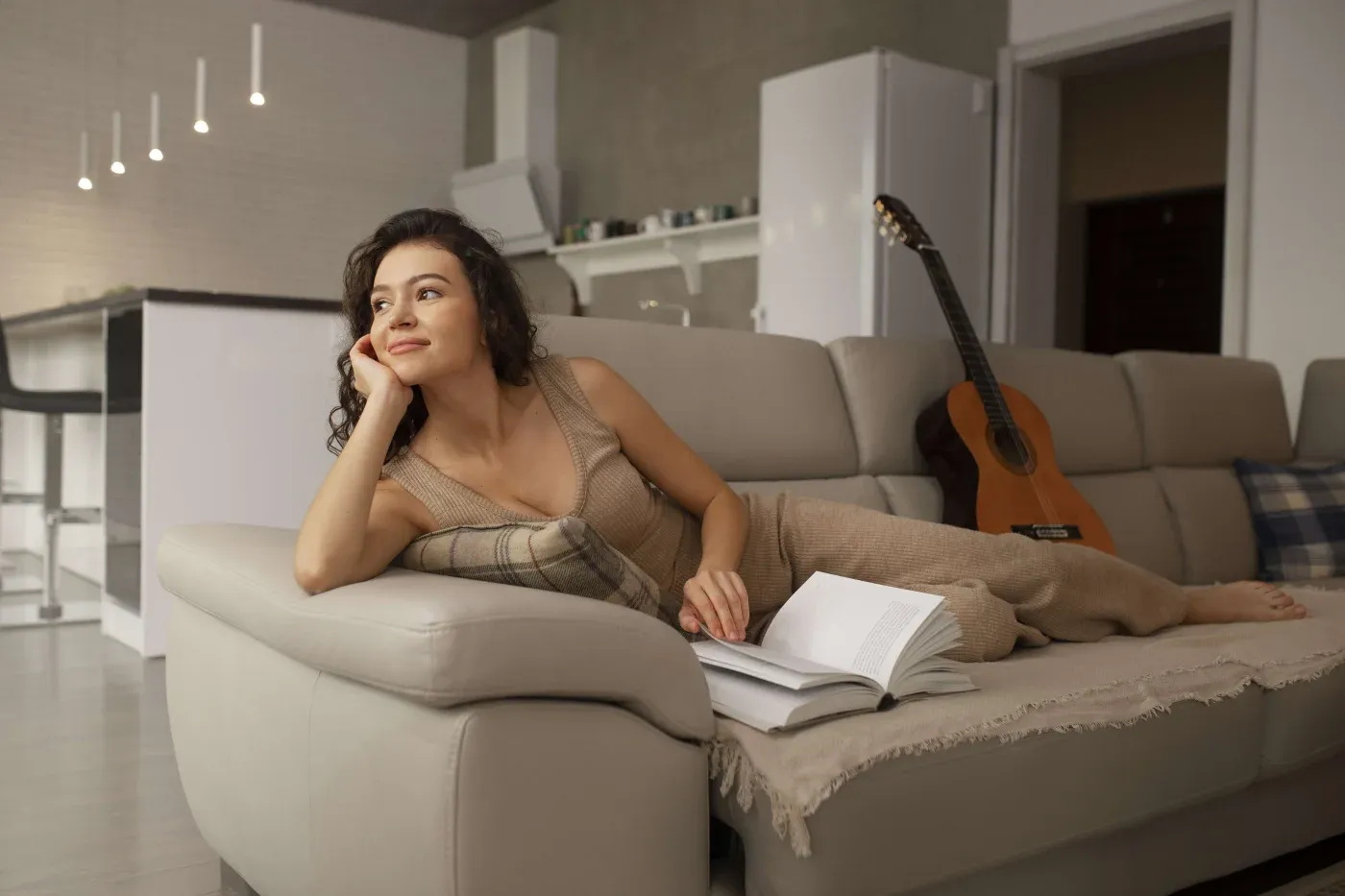 A woman in a cozy living room leans on a couch, gazing thoughtfully into the distance. An open book lies beside her, and a guitar rests against the couch, creating a relaxed and contemplative atmosphere.
