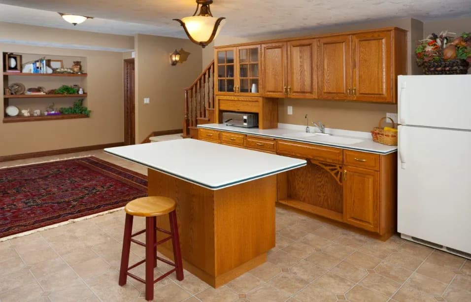 Warm kitchen with oak cabinets, a white countertop island, red stool, and adjacent living area with a red rug and wooden shelves. Cozy and inviting.