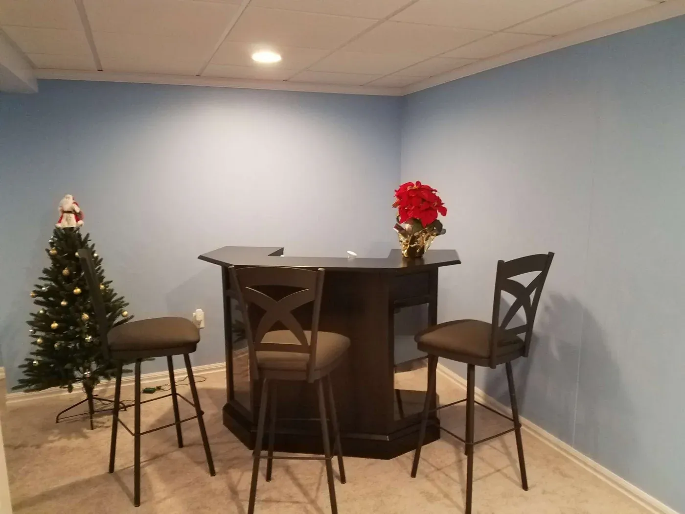 A home bar with three tall chairs in a corner of a room with light blue walls. A poinsettia decorates the bar; a small Christmas tree stands nearby.