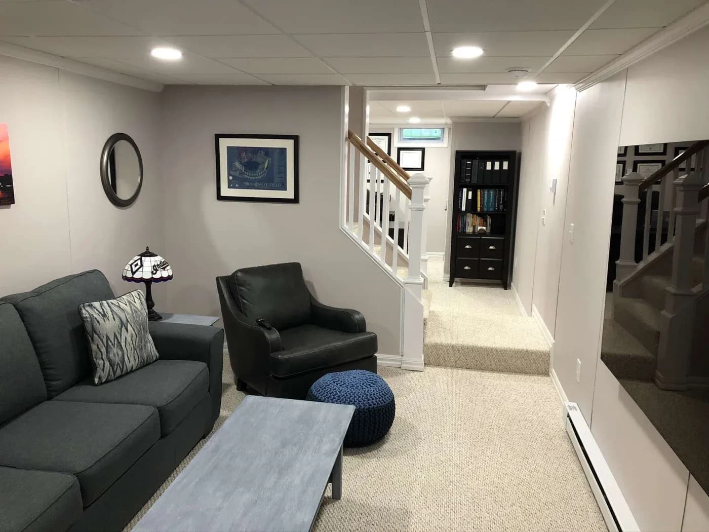 Cozy basement lounge with a gray sofa, armchair, and blue ottoman. A small table and lamp sit nearby. Stairs and bookshelf in the background.
