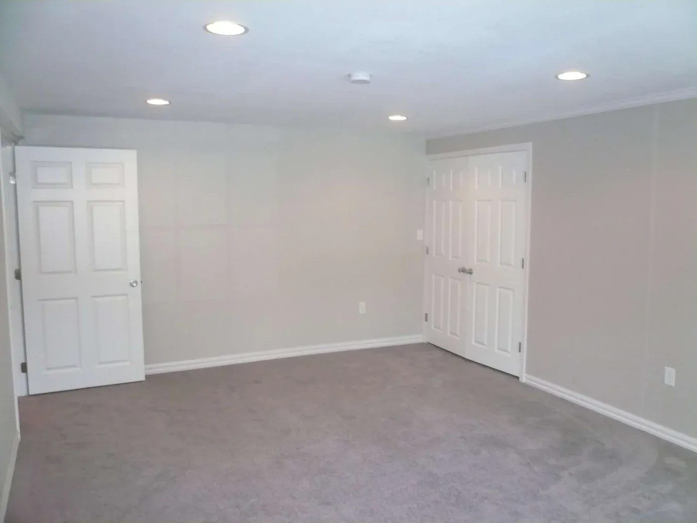 Spacious empty room with beige carpet, light gray walls, recessed ceiling lights, a single door on the left, and double doors on the right.