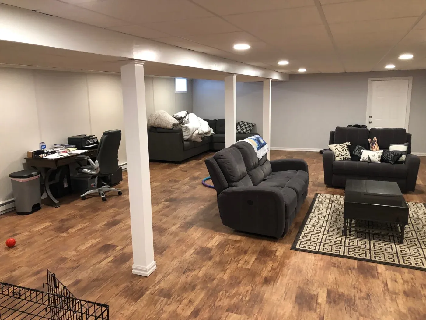 Spacious basement with wood flooring, featuring a cozy living area with gray sofas and cushions, a patterned rug, and a small office desk setup.