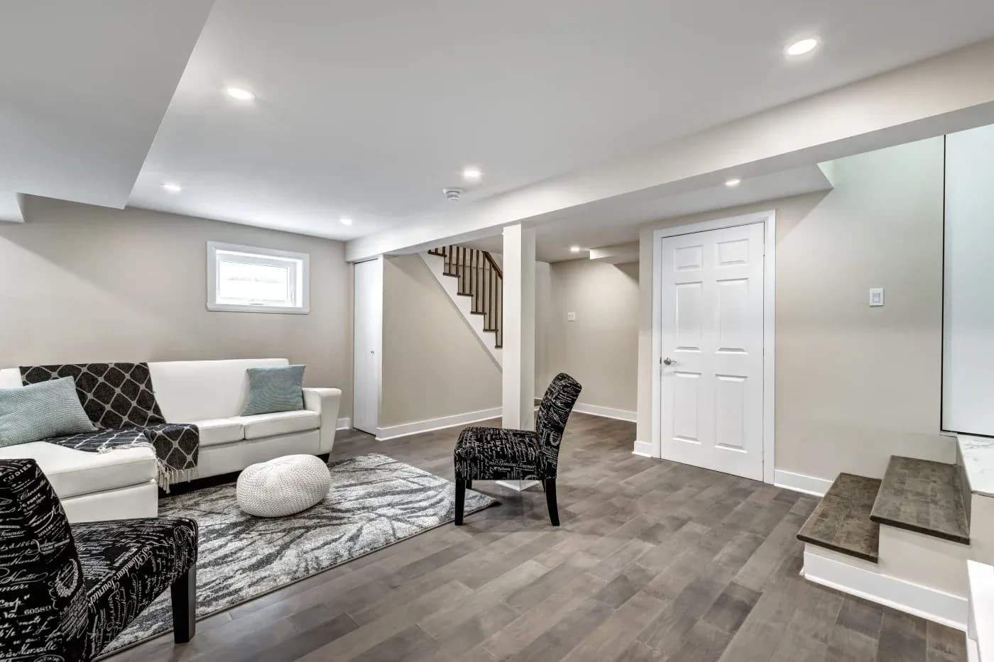 Modern basement with beige walls, recessed lighting, and dark wood flooring. Features a white sofa, patterned chairs, rug, pouf, and stairway. Cozy and inviting.