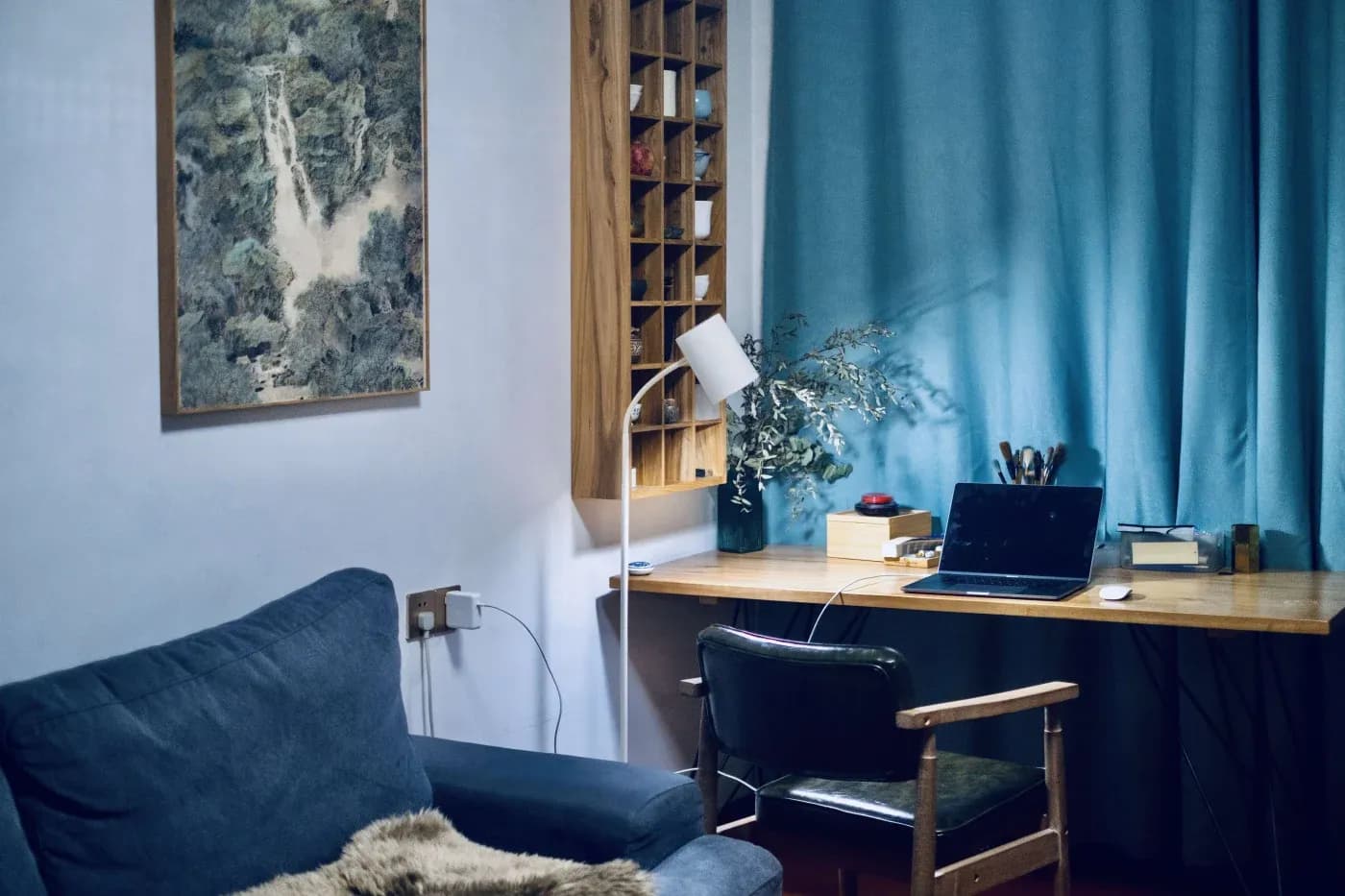 Cozy home office with a wooden desk, laptop, and books. A blue curtain and gray armchair enhance the tranquil atmosphere. Wall art adds interest.