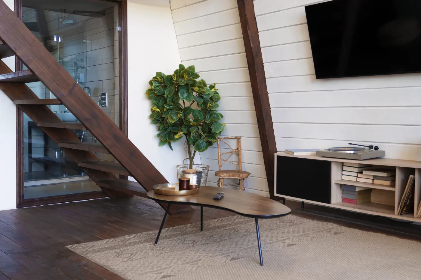 A modern living room features a wooden staircase, a large leafy plant, a triangular coffee table, and a wall-mounted TV, creating a cozy atmosphere.