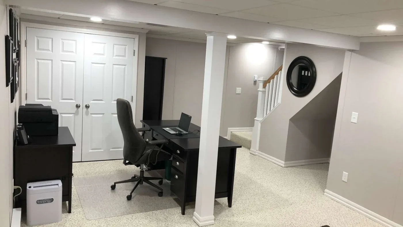A clean, modern home office in a basement features a desk with a laptop and office chair, beige carpet, a printer, and a staircase with white railings.
