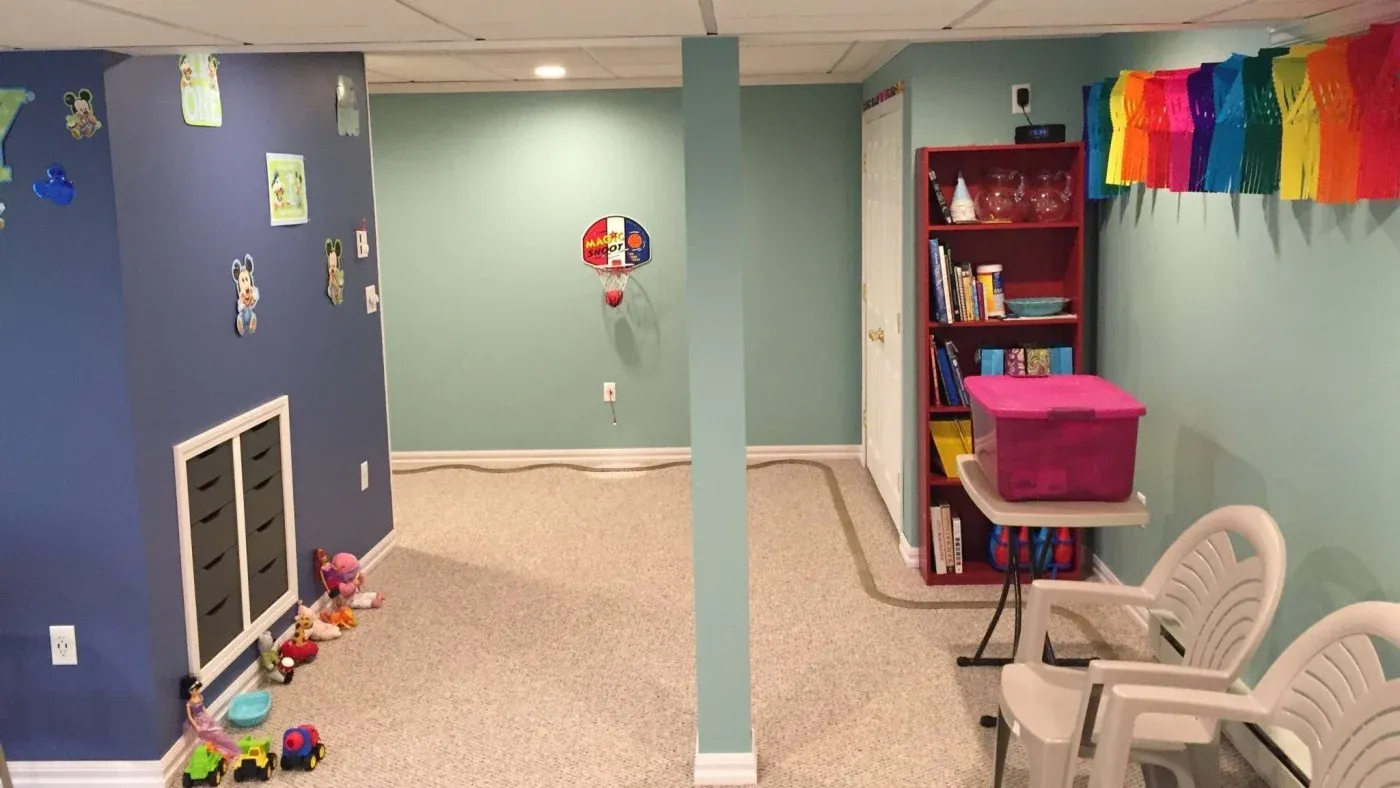 A colorful children's playroom with blue and teal walls, toys lining the floor, a hoop on the wall, a red bookshelf with books, and a rainbow banner.