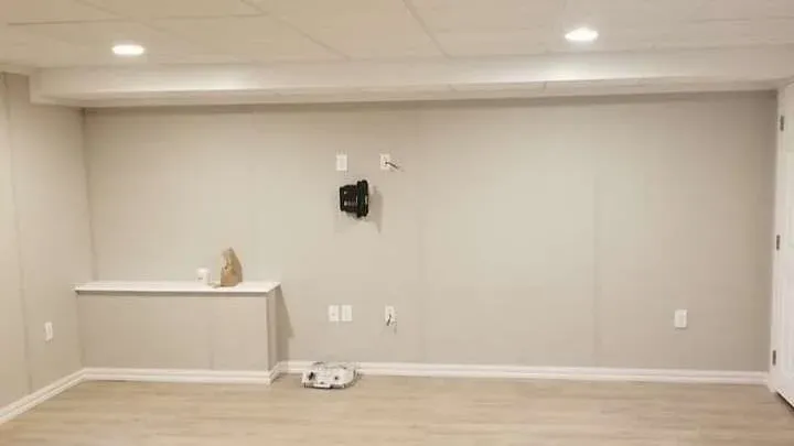 Minimalistic gray basement with light wood flooring, recessed lighting, and wall outlets. A shelf holds a paper bag and mug, creating a calm, empty space.