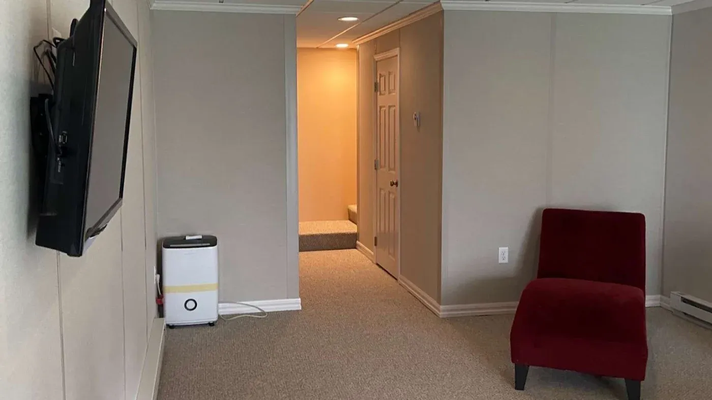 A minimalist room features a mounted TV on the left, a red chair on the right, beige walls, a small white appliance, and a lit hallway in the background.