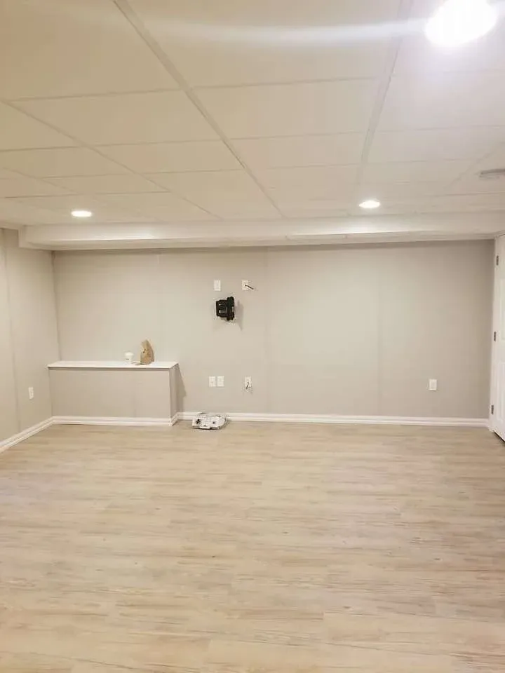 An empty basement with light wood flooring, beige walls, and recessed ceiling lights. A TV mount is on the wall. The space feels clean and modern.