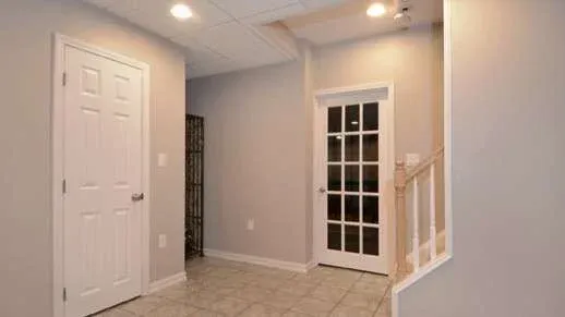 A clean, empty hallway features a closed white door on the left, a glass-paneled door straight ahead, soft gray walls, and tiled flooring, conveying a modern, minimalistic vibe.