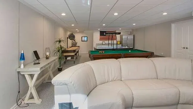 A cozy basement features a large white sectional sofa, pool table, and American flag on the wall. Bright lighting and modern decor create a relaxed vibe.
