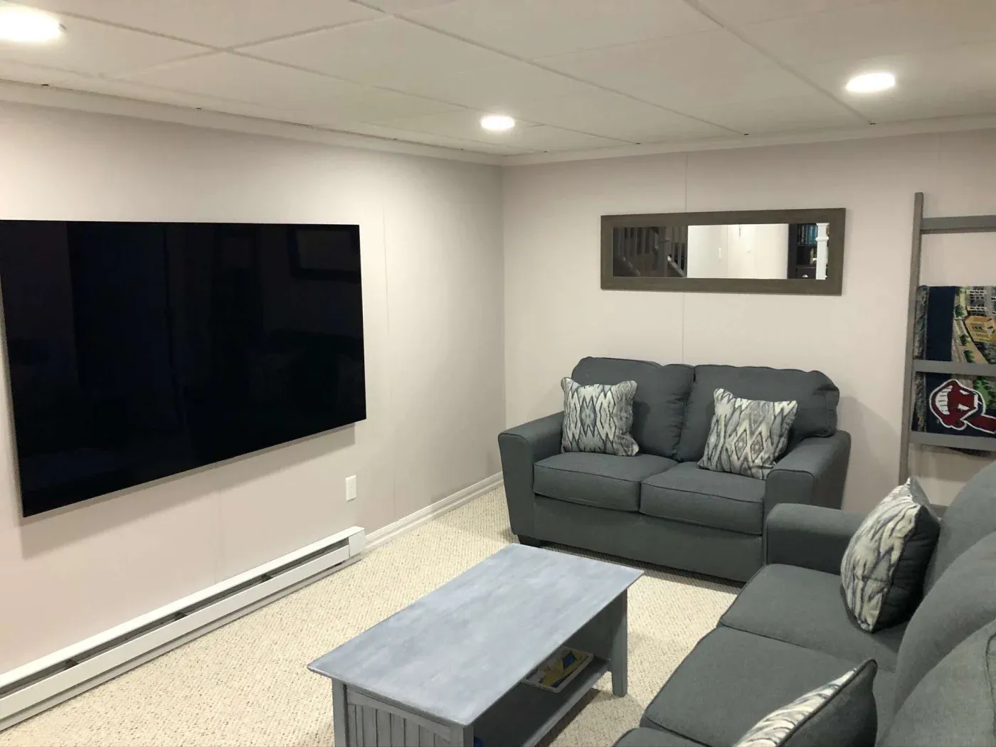 A cozy basement living room with gray sofas adorned with patterned cushions. A large flat-screen TV is mounted on the left wall. A wooden coffee table and a wall mirror complete the setting.