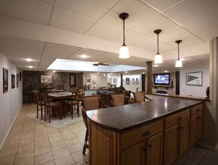 Spacious basement with a cozy ambiance features a bar area with wooden cabinets and hanging lights, a dining table, sofas, and a TV on a stone wall.