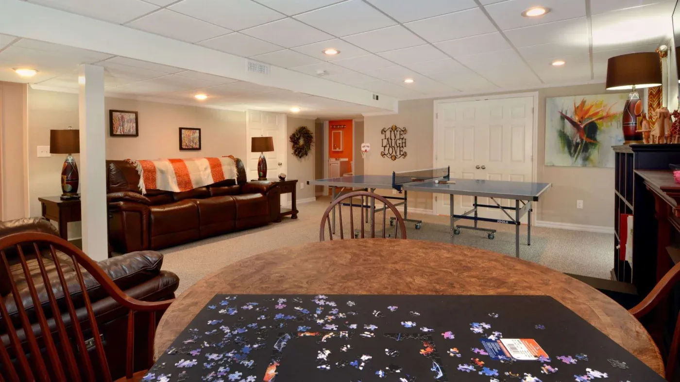 A cozy basement with a warm ambiance featuring a puzzle on a wooden table, a leather recliner sofa, colorful wall art, and a ping pong table.