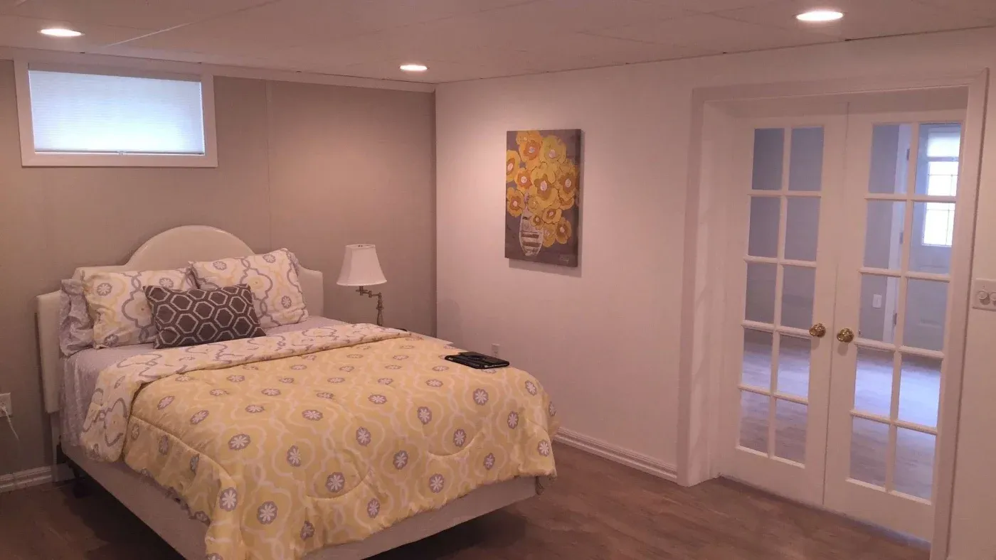 A cozy bedroom with a soft, warm ambiance. Features a bed with a yellow floral comforter, patterned pillows, a bedside lamp, floral artwork, and French doors.