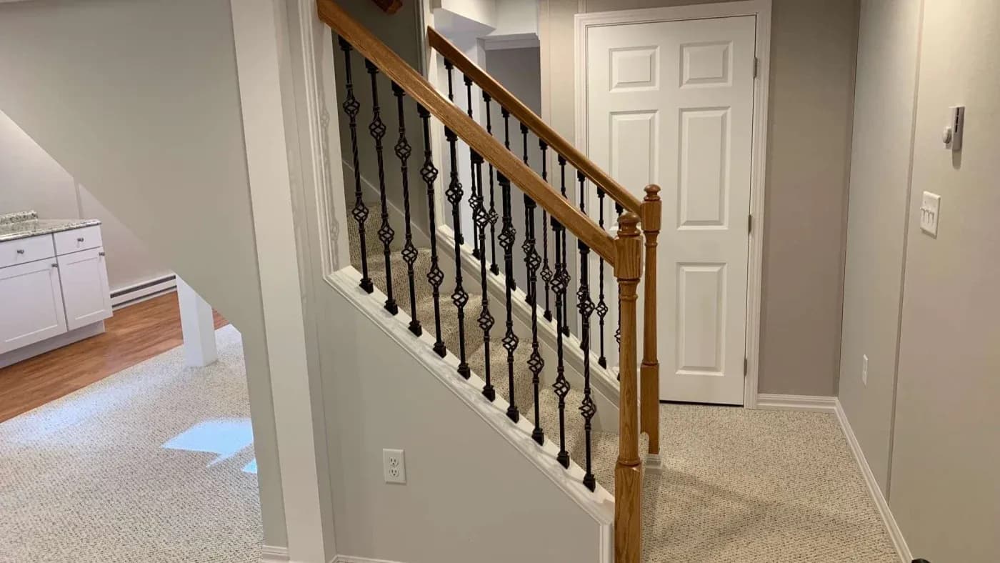 A cozy interior showcases a staircase with wooden handrail and decorative black iron balusters. Nearby, a closed white door and carpeted flooring.