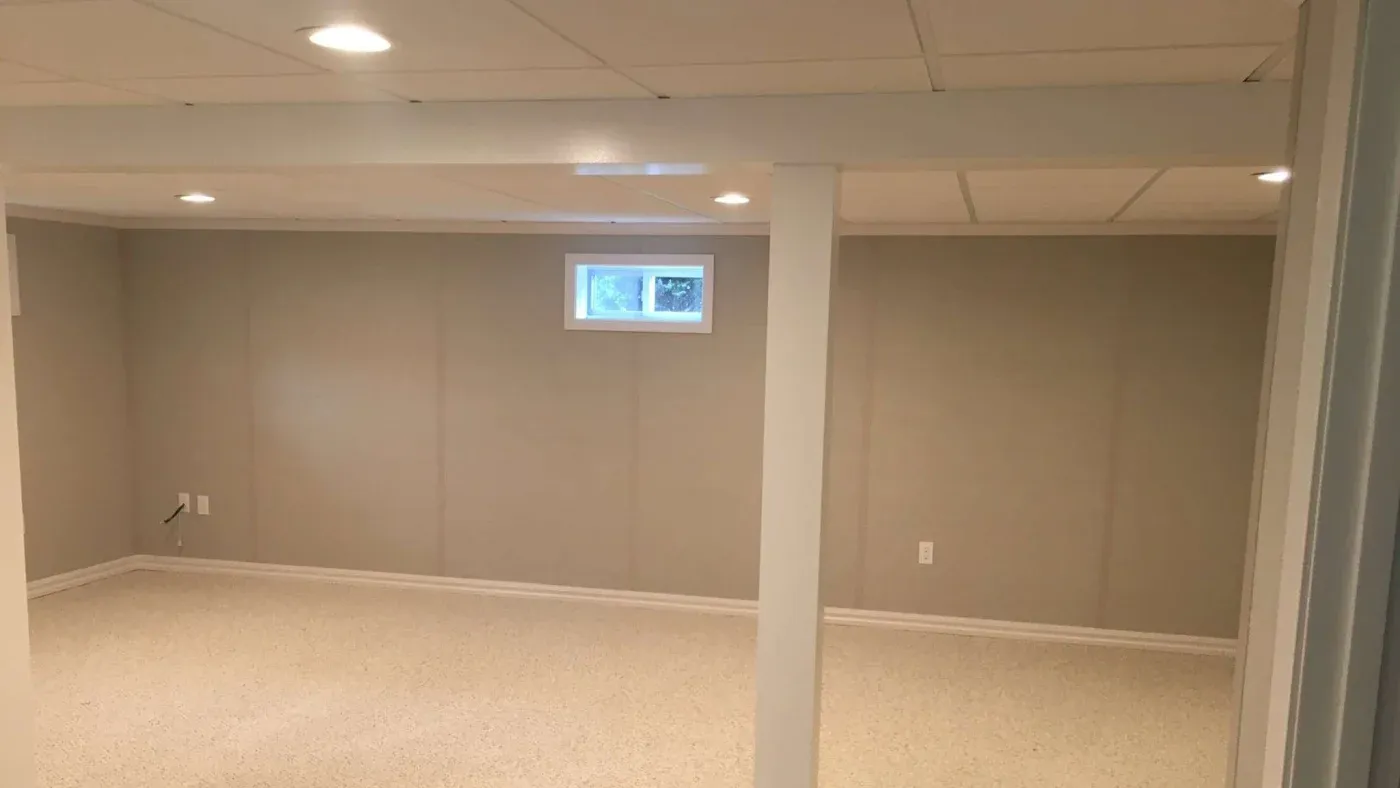 A basement room with beige walls, off-white carpet, a small rectangular window, recessed lighting, and support beams, creating a clean, spacious feel.