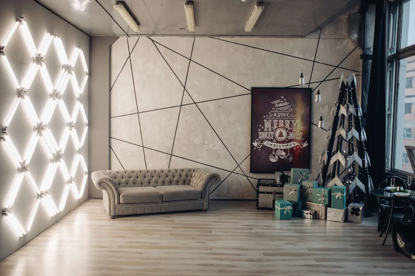 Modern living room with a geometric wall, velvet sofa, and bright light fixtures. A decorated Christmas tree with gifts and a festive sign create a cozy holiday vibe.
