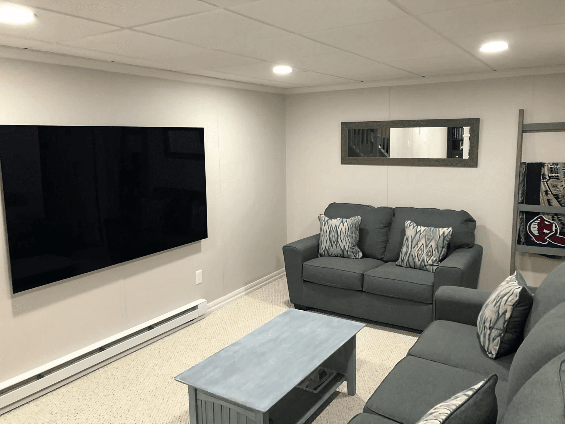 Cozy living room with gray couches, patterned cushions, and a large wall-mounted TV. A blue coffee table and wall mirror add to the relaxed ambiance.