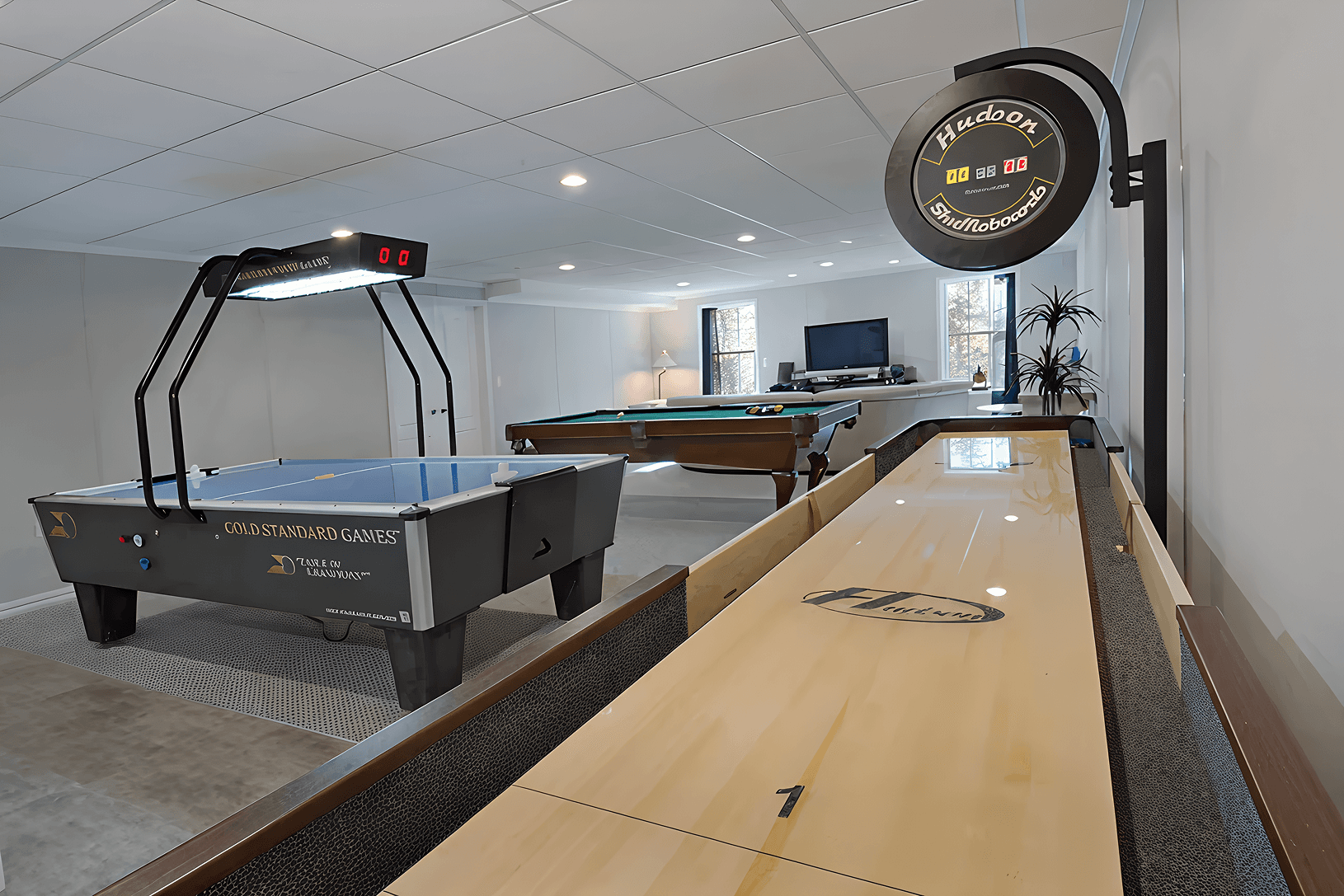 A spacious game room featuring a shuffleboard table in the foreground, an air hockey table, a pool table, and a TV. The atmosphere is bright and inviting.