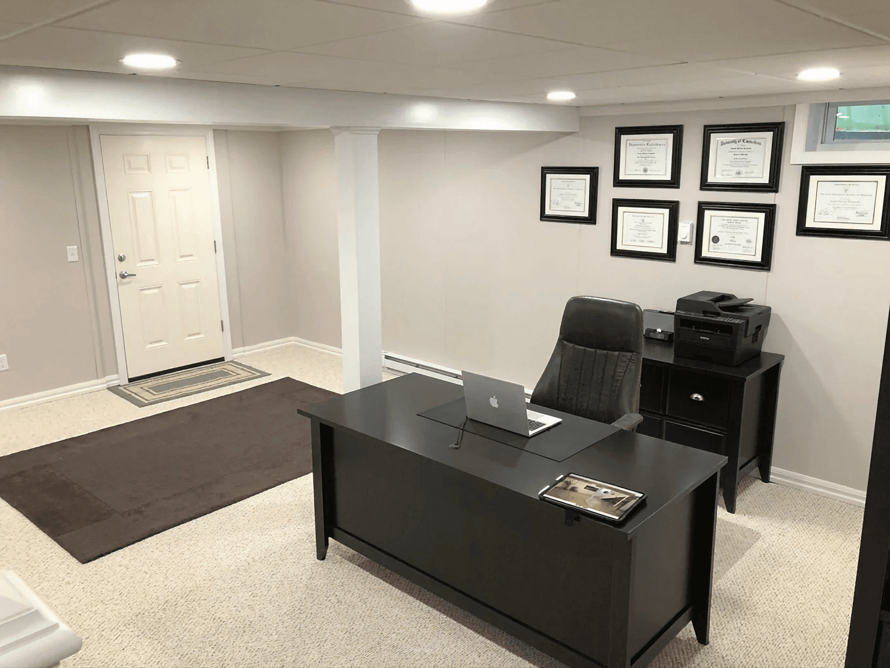 A tidy, well-lit office with a dark desk, a laptop, and a chair. Certificates are framed on the wall, and a printer sits on a side table. Calm and professional atmosphere.