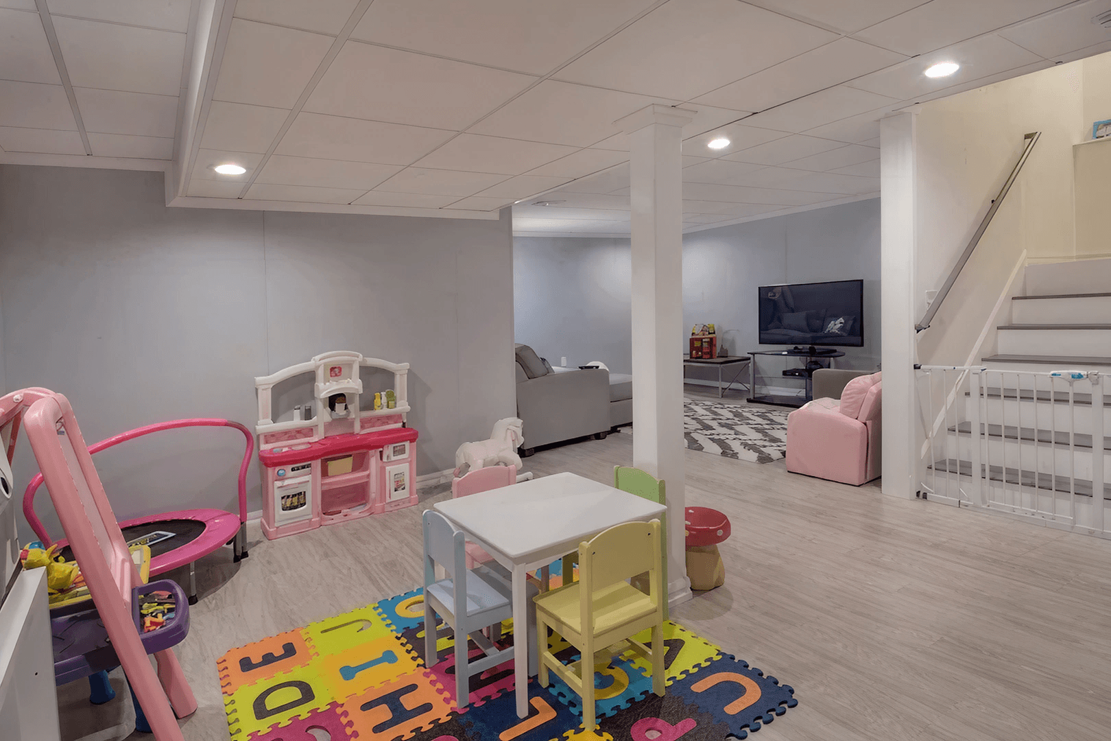 Spacious playroom with colorful foam mats, toy kitchen, and table set. Cozy seating area with TV in the background. Bright, welcoming atmosphere.